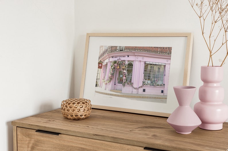 May include: A framed print of a pink storefront with a sign that reads "MERRY PUCKLECHURCH Indies". The storefront is decorated with flowers and greenery. The print is on a wooden table with a wicker basket and two pink vases.