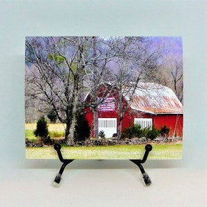 May include: A print of a red barn with white doors and a rusty roof, set against a backdrop of trees and a field. The artwork is displayed on a black metal easel.