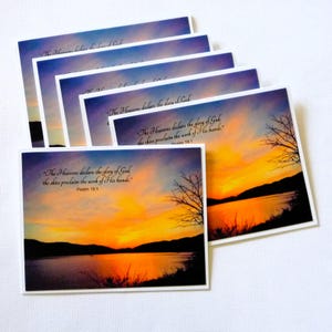 May include: A stack of greeting cards featuring a sunset over a lake. The sky transitions from blue to orange and yellow. The cards have the text "The Heavens declare the glory of God, the skies proclaim the work of His hands." Psalm 19:1.
