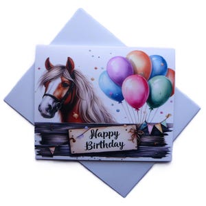 May include: A birthday card featuring a horse head with a white mane, balloons, and the text "Happy Birthday." The balloons are in various colors, including pink, purple, and blue. The card is on a white background with a light blue envelope.