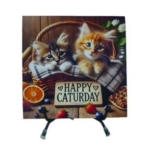 May include: A decorative tile featuring two kittens in a basket with a "Happy Caturday" sign. The kittens are orange and brown, with blue eyes. The tile is supported by a black metal stand.