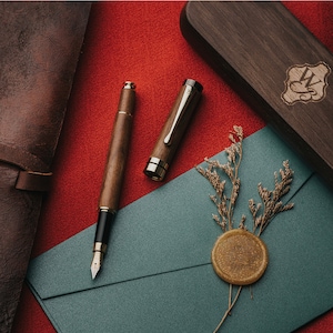 Op de afbeelding: Twee houten pennen met gouden accenten en zwarte puntjes, een in een houten doos en de andere op een groene envelop met een gouden zegel van was en gedroogde bloemen.