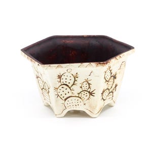 May include: A cream-colored, hexagonal ceramic planter with a dark brown interior. The exterior features a hand-drawn cactus design. The planter has a distressed, vintage look and is elevated by small feet.