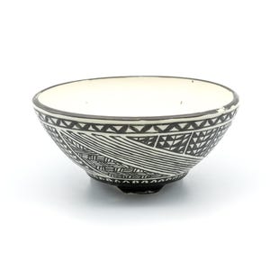 Può includere: Una piccola ciotola in ceramica con interno bianco e un motivo geometrico nero all'esterno. Il design della ciotola presenta una serie di linee, triangoli e altre forme. Il bordo è scuro e la ciotola poggia su una superficie bianca.