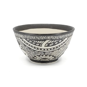 Può includere: Una ciotola in ceramica con bordo nero e interno bianco. L'esterno presenta un motivo geometrico in bianco e nero. La ciotola è rotonda e poggia su una superficie bianca. Il design è simmetrico e dettagliato.