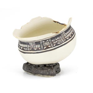 Può includere: Ciotola in ceramica fatta a mano, di colore crema, con un motivo geometrico nero sul bordo. La ciotola poggia su una base scura e strutturata. Il bordo ha una forma irregolare e organica.