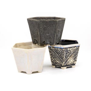 May include: Three small, square ceramic planters in a stack. The top planter is dark gray with a textured surface. The middle planter is white with a glossy finish. The bottom planter is black and white with a geometric design.