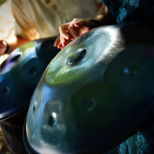 May include: Two steel handpan drums, one blue and one green, are held by two people. The drums are played by tapping the surface with fingers.