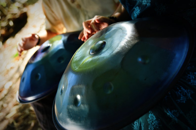 May include: Two steel handpan drums, one blue and one green, are held by two people. The drums are played by tapping the surface with fingers.