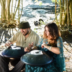 May include: Two people are sitting on the ground playing handpan drums. The handpans are dark gray and have a metallic finish. The people are wearing casual clothing and are smiling. The background is a bamboo forest.