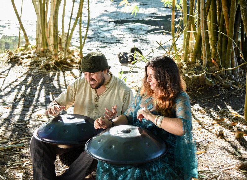 May include: Two people are sitting on the ground playing handpan drums. The handpans are dark gray and have a metallic finish. The people are wearing casual clothing and are smiling. The background is a bamboo forest.