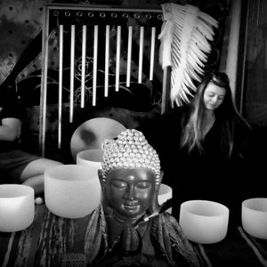 May include: A black and white photo of a person playing a flute and another person sitting with their eyes closed. There are several crystal singing bowls in the foreground, and a statue of a Buddha in the center.