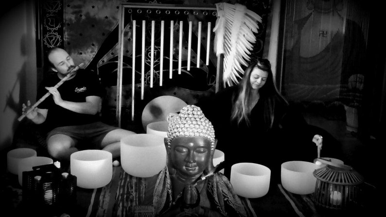 May include: A black and white photo of a person playing a flute and another person sitting with their eyes closed. There are several crystal singing bowls in the foreground, and a statue of a Buddha in the center.