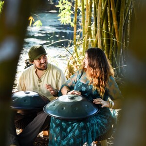 May include: Two people sitting outdoors playing handpan drums. The person on the left is wearing a beige shirt and a green hat. The person on the right is wearing a teal dress with a floral pattern. Both handpans are black.