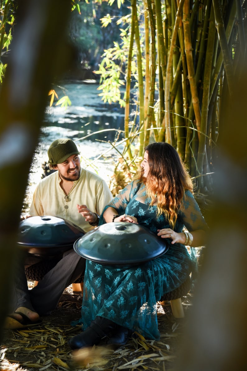 May include: Two people sitting outdoors playing handpan drums. The person on the left is wearing a beige shirt and a green hat. The person on the right is wearing a teal dress with a floral pattern. Both handpans are black.