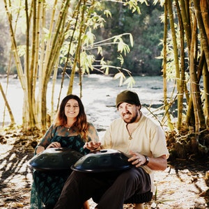 May include: Two people are sitting in a bamboo forest playing handpan drums. The handpans are black and round. The people are wearing casual clothing. The forest is green and lush.
