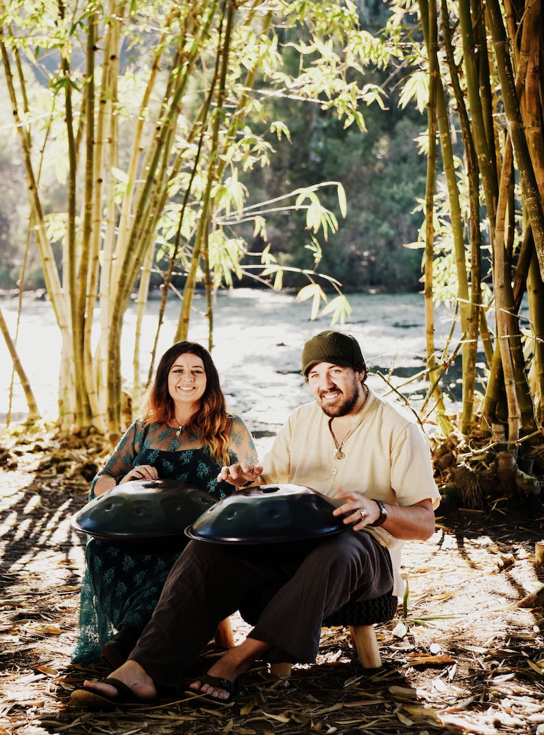 May include: Two people are sitting in a bamboo forest playing handpan drums. The handpans are black and round. The people are wearing casual clothing. The forest is green and lush.