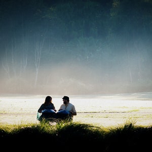 May include: Two people sit facing each other on a grassy field, with a misty forest in the background. They are playing a musical instrument that looks like a set of steel drums.