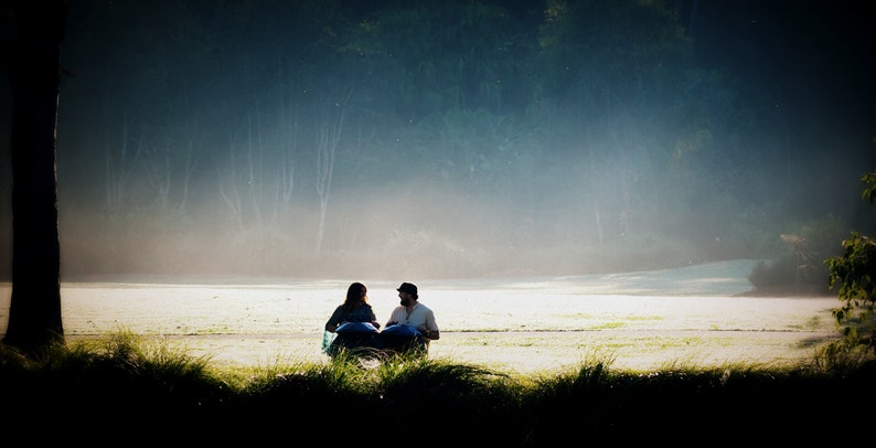 May include: Two people sit facing each other on a grassy field, with a misty forest in the background. They are playing a musical instrument that looks like a set of steel drums.