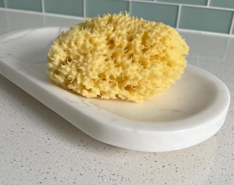 May include: A natural yellow sea sponge rests on a white marble soap dish. The dish has a smooth, oval shape. The background features a white and blue tiled wall.