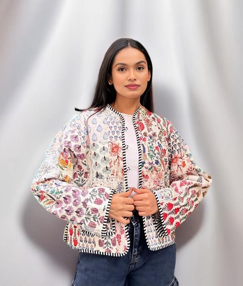 May include: A white quilted jacket with a floral patchwork design. The jacket has a mandarin collar, long sleeves, and a button closure. The jacket is trimmed with black piping.