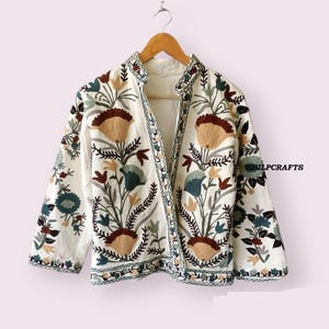 以下が含まれることがあります： 茶色、緑、青の色合いの花柄刺繍が施された白い綿のジャケット。ジャケットはスタンドカラーで、フロントに留め具があります。ジャケットの右側に「SHILPCRAFTS」という文字が見えます。