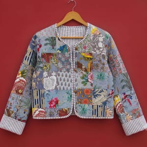 Puede incluir: Una chaqueta acolchada con un diseño de patchwork de varios patrones florales y botánicos en tonos azul, gris y blanco. La prenda tiene mangas largas con puños a rayas, cuello redondo y cierre frontal de botones con un borde a rayas.