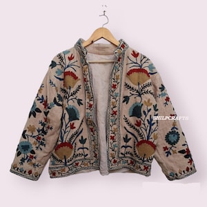 Può includere: Una giacca beige ricamata con un motivo floreale in tonalità di blu, rosso e oro. La giacca ha un colletto alla coreana e una chiusura con cerniera sul davanti. Il testo "SHILPCRAFTS" è visibile sul lato destro della giacca.