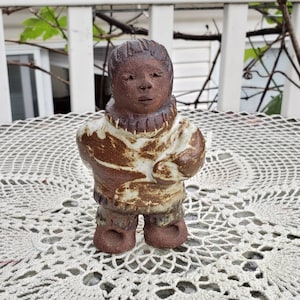 May include: A hand-sculpted ceramic figurine of a person wearing a textured parka with a high collar. The figure features a dark brown clay finish with mottled white glaze accents on the torso and sleeves, showcasing a rustic, earthy aesthetic.