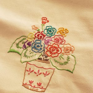 May include: Embroidered floral design featuring a vase filled with colorful flowers. The flowers are in shades of red, blue, yellow, and pink, with green leaves. The vase is a light peach color with a decorative pattern.
