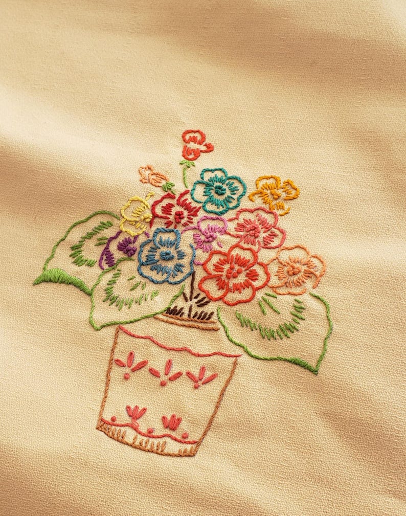 May include: Embroidered floral design featuring a vase filled with colorful flowers. The flowers are in shades of red, blue, yellow, and pink, with green leaves. The vase is a light peach color with a decorative pattern.