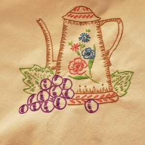 May include: Embroidered cream-colored teapot with a floral design in pink, blue, and green. A cluster of purple grapes and green leaves are embroidered below the teapot. The teapot has a brown outline.