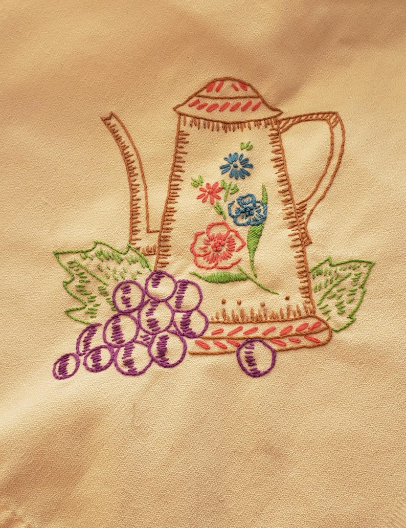 May include: Embroidered cream-colored teapot with a floral design in pink, blue, and green. A cluster of purple grapes and green leaves are embroidered below the teapot. The teapot has a brown outline.