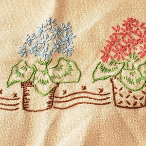May include: Embroidered floral design featuring three potted flowers in shades of peach, blue, and pink. The flowers have green leaves and are set against a cream-colored background. The pots are brown and sit above a brown wavy line.