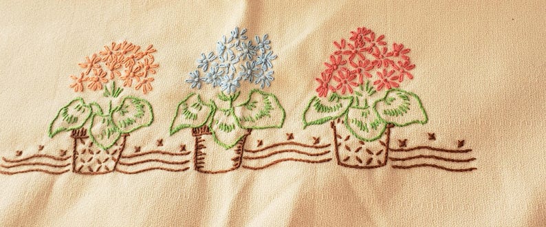 May include: Embroidered floral design featuring three potted flowers in shades of peach, blue, and pink. The flowers have green leaves and are set against a cream-colored background. The pots are brown and sit above a brown wavy line.