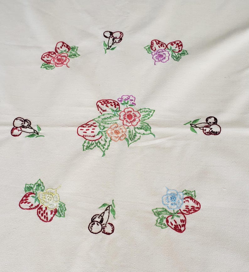 May include: Cream-colored fabric with embroidered designs of strawberries, cherries, and flowers. The embroidery features red, pink, purple, and green colors. The arrangement of the embroidered elements is scattered across the fabric.