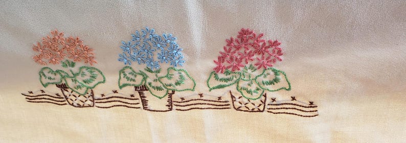 May include: Embroidered floral design on a cream-colored fabric. Three flower pots with pink, blue, and coral flowers are displayed. The flowers are above green leaves and brown woven baskets. A brown stitched line runs below the baskets.