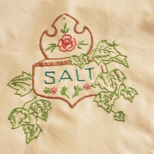 May include: Embroidered cream-colored fabric with a brown outline of a decorative salt container. The word "SALT" is embroidered in teal, with green leaves and pink flowers. A vintage kitchen textile.