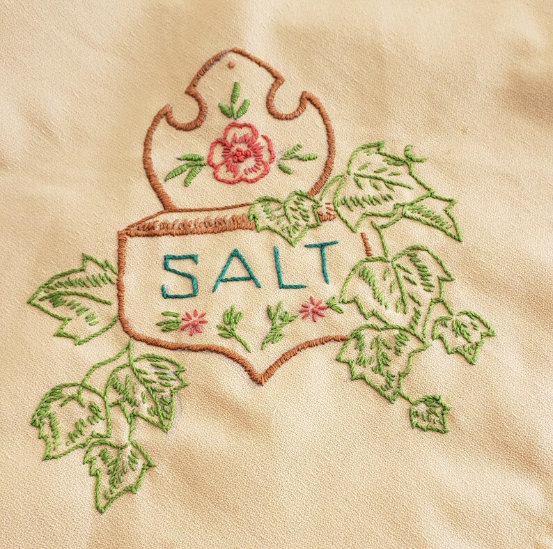 May include: Embroidered cream-colored fabric with a brown outline of a decorative salt container. The word "SALT" is embroidered in teal, with green leaves and pink flowers. A vintage kitchen textile.