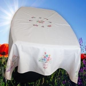 May include: White tablecloth with embroidered floral patterns. The tablecloth has colourful embroidered flowers and a watering can design on the edges. The flowers are in shades of pink, blue, and green. The tablecloth is draped over a table.