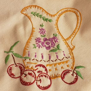 May include: Embroidered pitcher design with cherries. The pitcher is cream-colored with an orange outline and handle. It features purple flowers and green leaves. Red cherries with green stems are at the bottom. The embroidery is on a cream-colored fabric.