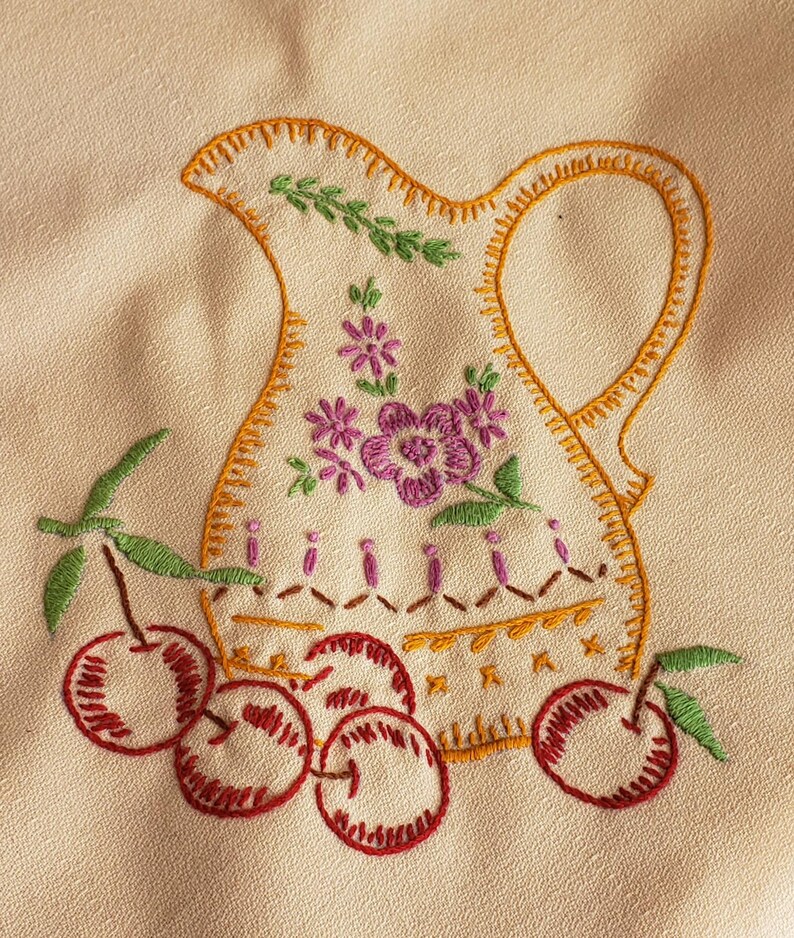 May include: Embroidered pitcher design with cherries. The pitcher is cream-colored with an orange outline and handle. It features purple flowers and green leaves. Red cherries with green stems are at the bottom. The embroidery is on a cream-colored fabric.