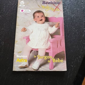 May include: A vintage book cover titled "Beehive Baby Talk" featuring a baby in a white dress and pink tights sitting on a pink chair. The cover has French text and a retro design.