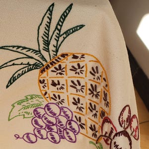 May include: Cream-colored fabric with embroidered fruit design. A yellow pineapple with brown details is next to a bunch of purple grapes and red cherries. Green leaves and stems complete the design. The embroidery is done in a detailed, vintage style.
