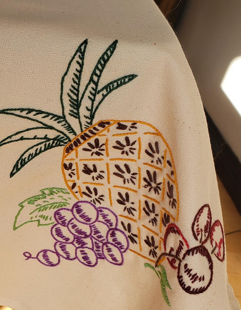 May include: Cream-colored fabric with embroidered fruit design. A yellow pineapple with brown details is next to a bunch of purple grapes and red cherries. Green leaves and stems complete the design. The embroidery is done in a detailed, vintage style.