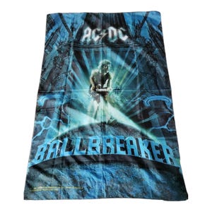 May include: A rectangular AC/DC Ballbreaker beach towel featuring a blue and black graphic print. The design displays a guitarist in a spotlight, surrounded by architectural elements and lightning, with the band name and album title text.