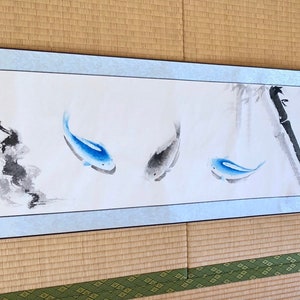 May include: A long, rectangular scroll featuring a watercolor painting of three fish and bamboo stalks. The fish are blue and gray, and the bamboo is black and gray. The scroll has a light blue border and is displayed on a woven mat.