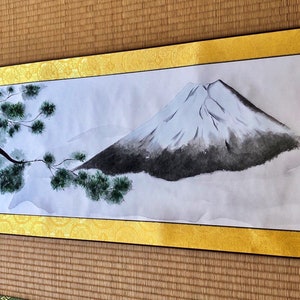 May include: Japanese-style scroll painting featuring Mount Fuji with a snow-capped peak, rendered in shades of gray and white. A tree with green foliage is on the left. The scroll has a gold border and is displayed on a woven mat.
