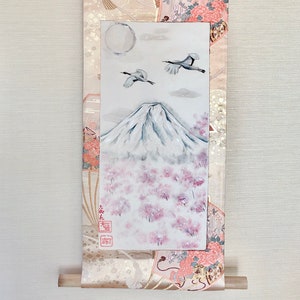 May include: Japanese wall scroll depicting Mount Fuji, cherry blossoms, and cranes. The scroll has a decorative border with floral and geometric patterns in pink, gold, and cream. The painting is framed by a patterned fabric.
