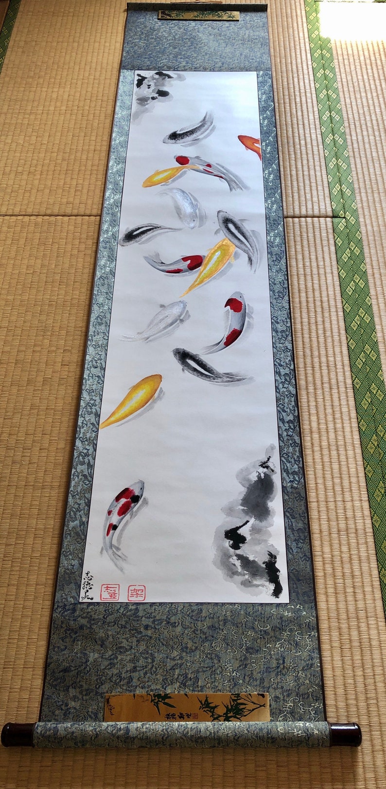 May include: A Japanese scroll painting featuring koi fish in shades of red, yellow, black, and white. The artwork is mounted on a scroll with a patterned border and wooden dowels.